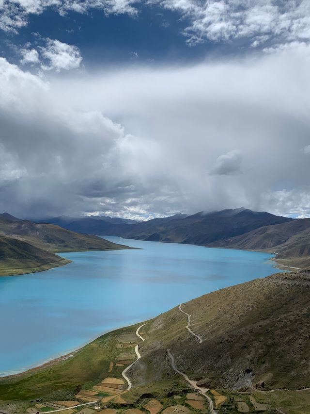A stunning turquoise lake surrounded by rugged hills under a dramatic cloudy sky in the Himalayas.